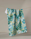 The Taryn Brennan "Leafy Bush" tea towel, ultra-absorbent and adorned with a green and yellow botanical leaf pattern, hangs on a line against a plain beige background.