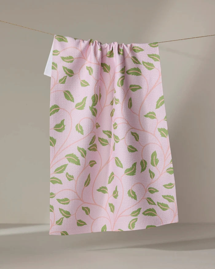 The Leafy Maze tea towel by Jasmine Blooms Designs features a pink fabric with green leaf patterns, made from recycled materials. Its subtly textured cloth is gathered at the top and hangs from a string against a neutral backdrop.