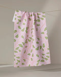 The Leafy Maze tea towel by Jasmine Blooms Designs features a pink fabric with green leaf patterns, made from recycled materials. Its subtly textured cloth is gathered at the top and hangs from a string against a neutral backdrop.
