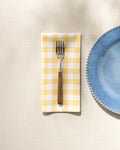 A fork rests on a GEOMETRY Lemon Gingham washable dinner napkin beside a blue textured-rim ceramic plate, all set on a cream-colored tablecloth.