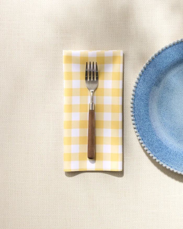 A fork rests on a GEOMETRY Lemon Gingham washable dinner napkin beside a blue textured-rim ceramic plate, all set on a cream-colored tablecloth.