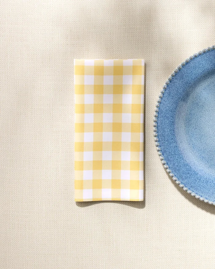 The GEOMETRY Lemon Gingham washable dinner napkin, in yellow and white checks, is neatly folded beside a blue plate with a textured rim on a light beige tablecloth.