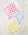 Ashley Celena’s Lemon Pop set features three patterned napkins—pink and white checks, blue with yellow lemons and red branches, and yellow with a white sunburst—crafted from recycled materials on a marble surface.