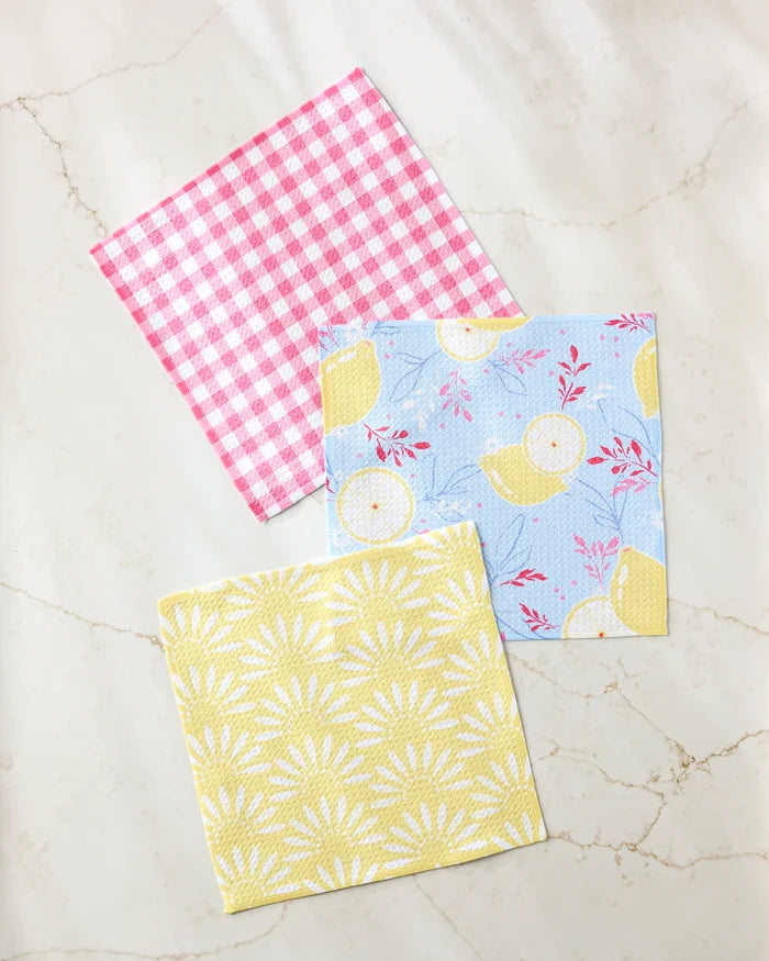 Ashley Celena’s Lemon Pop set features three patterned napkins—pink and white checks, blue with yellow lemons and red branches, and yellow with a white sunburst—crafted from recycled materials on a marble surface.