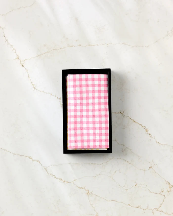 The Ashley Celena Lemon Pop box with a pink and white checkered lid sits on a white marble surface with tan veins, ideal for storing reusable paper towels in your eco-friendly kitchen.