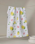 Lemons in Capri" by Travel Write Draw features a white tea towel with lemons, leaves, and red swirls—made from recycled materials—hanging above stacked plates against a neutral background.