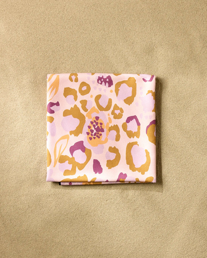 The Leopard Loving silk scarf by Elisabeth Wing, made from recycled materials and featuring a pink, yellow, and purple abstract animal print, rests on a textured beige surface.
