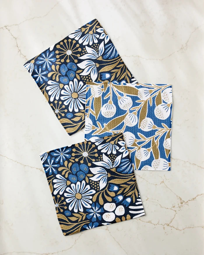 Leslie Flowers by Amy MacCready: three patterned cloth napkins in blue, white, and gold with floral and botanical designs are displayed on a marble surface—part of an eco-friendly set made from recycled materials.