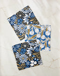 Leslie Flowers by Amy MacCready: three patterned cloth napkins in blue, white, and gold with floral and botanical designs are displayed on a marble surface—part of an eco-friendly set made from recycled materials.