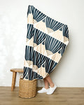 A person holds up the Amy MacCready Letitia Dark blanket—featuring bold black and beige patterns—obscuring their upper body, with only their legs in white sneakers visible next to a stool and woven basket on a wooden floor.