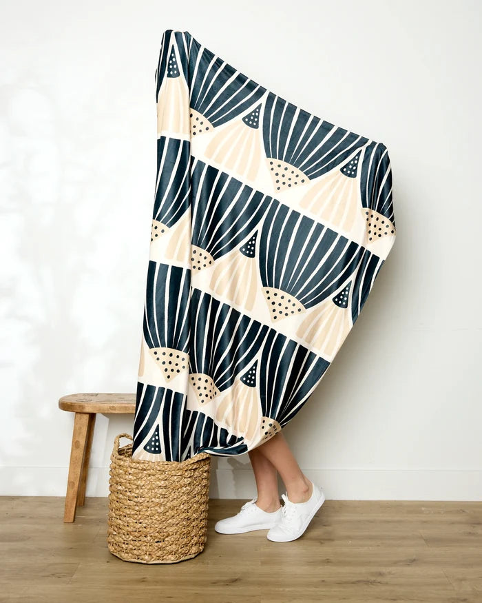 A person holds up the Amy MacCready Letitia Dark blanket—featuring bold black and beige patterns—obscuring their upper body, with only their legs in white sneakers visible next to a stool and woven basket on a wooden floor.