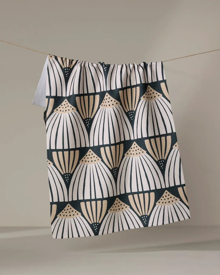 The Letitia recycled kitchen towel by Amy MacCready features rows of stylized fan or shell shapes in a geometric pattern with black, white, and tan semicircles, and hangs from a string against a neutral background.