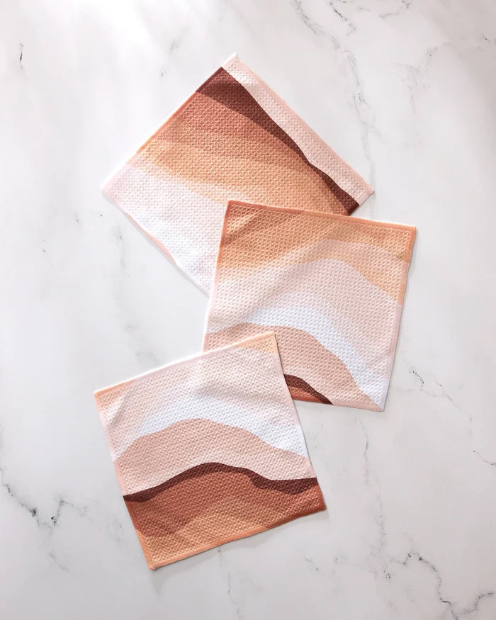 Three GEOMETRY "Levels of Fall" cloths, made from recycled materials and featuring abstract wavy patterns in brown, beige, and pink tones, are displayed on a white marble surface.