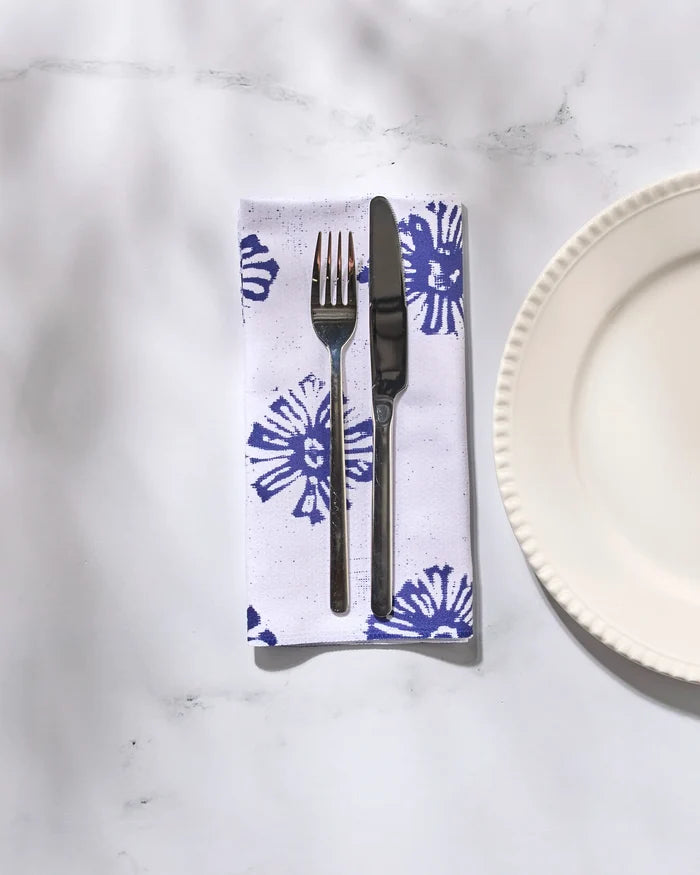A fork and knife rest on a folded reusable Geometry Leyla White cloth napkin with blue floral designs beside a white, scalloped-edge plate on a marble surface.