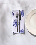 A fork and knife rest on a folded reusable Geometry Leyla White cloth napkin with blue floral designs beside a white, scalloped-edge plate on a marble surface.