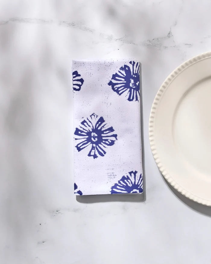 The Geometry Leyla White reusable cloth napkin, featuring a blue floral pattern, is neatly folded beside a white scalloped plate on marble—an elegant addition to any dinner napkin set.