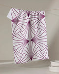 The Amy MacCready Libby Dark Maroon kitchen tea towel, made from recycled materials and ultra-absorbent, hangs on a thin string. Its dark maroon geometric fan pattern stands out with a stack of white plates in the neutral background.