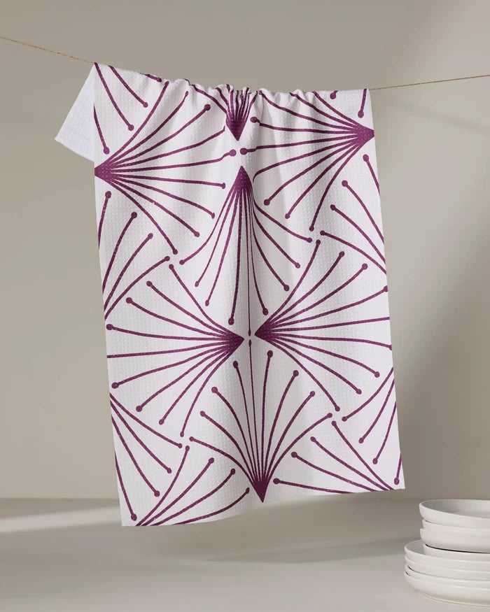The Amy MacCready Libby Dark Maroon kitchen tea towel, made from recycled materials and ultra-absorbent, hangs on a thin string. Its dark maroon geometric fan pattern stands out with a stack of white plates in the neutral background.