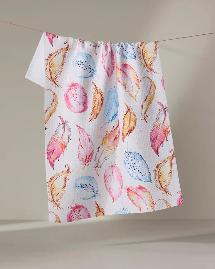 The Elena Fay "Light as a Feather" towel, ultra-absorbent and white with a vibrant feather pattern in pink, orange, yellow, and blue, is displayed hanging against a plain light background.