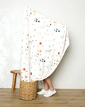A person stands in a woven basket, wrapped mostly in the Coit Creative Light Farm Animals blanket with animal and plant patterns. Only their lower legs and white shoes are visible. A wooden stool rests nearby against a plain wall and wood floor.