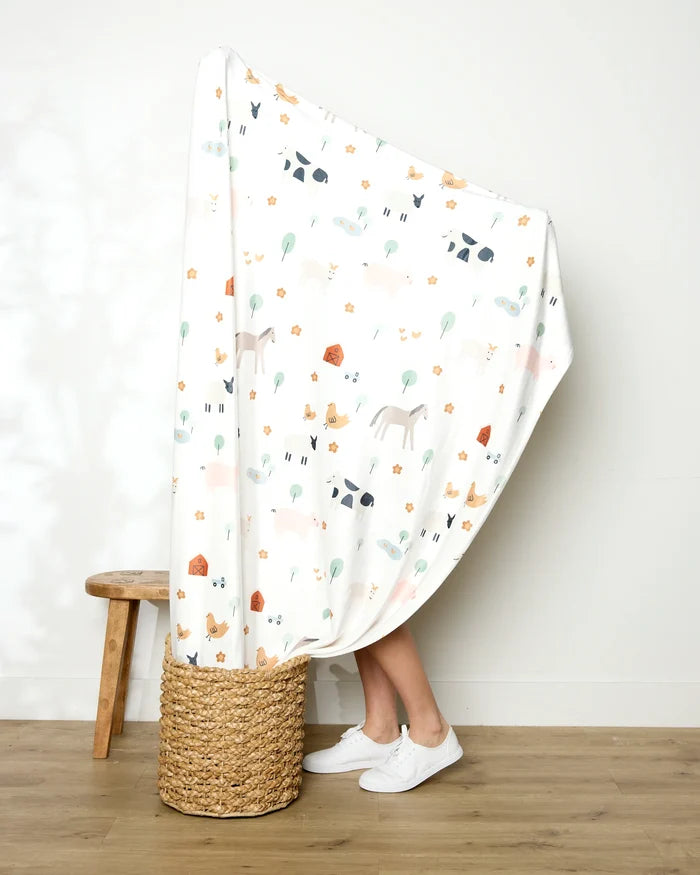 A person stands in a woven basket, wrapped mostly in the Coit Creative Light Farm Animals blanket with animal and plant patterns. Only their lower legs and white shoes are visible. A wooden stool rests nearby against a plain wall and wood floor.