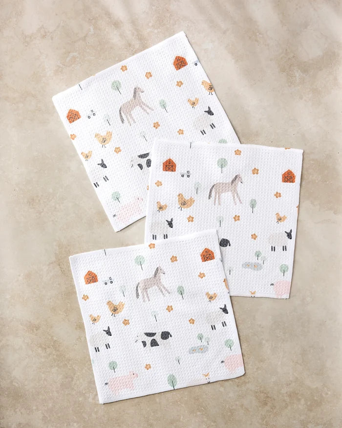 Three white paper napkins from Coit Creative's Light Farm Animals collection, featuring horses, cows, chickens, pigs, trees, flowers & barns on a beige surface—perfect paired with recycled dish towels for any farmhouse kitchen.