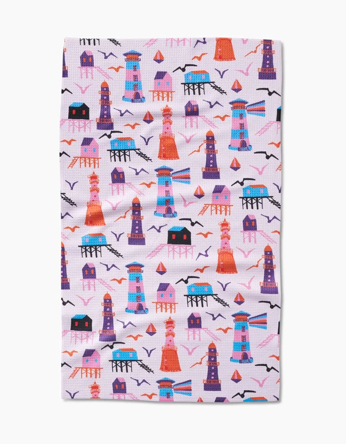 The Lighthouse In Pink tea towel by Ania Zwara Art features lighthouses, houses, seagulls, and boats in vibrant colors on a light background. Made from ultra-absorbent Geoweave fabric for style and functionality.