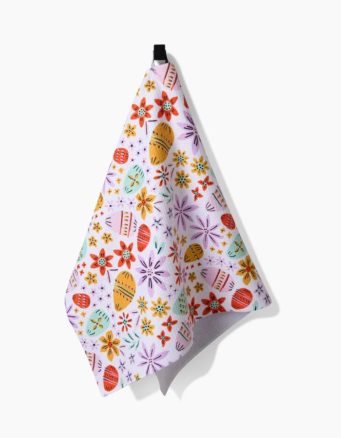 The Lilith tea towel by Amy MacCready features a colorful Easter egg and flower pattern in red, orange, yellow, purple, and teal on white recycled fabric. It hangs from a black loop against a white background.
