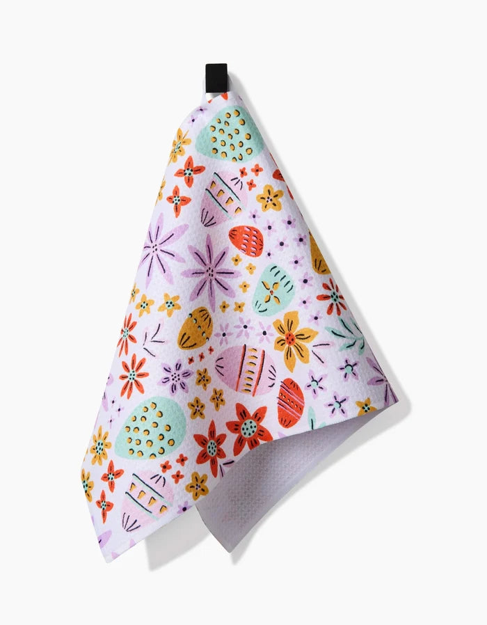 The Lilith bar towel by Amy MacCready is ultra-absorbent and white, featuring colorful flower and Easter egg patterns. It hangs from a black loop, shown against a white background.