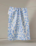 The Geometry Lilou Cream kitchen towel, made from recycled materials and featuring a textured surface with a blue floral pattern, hangs on a string against a light gray background.