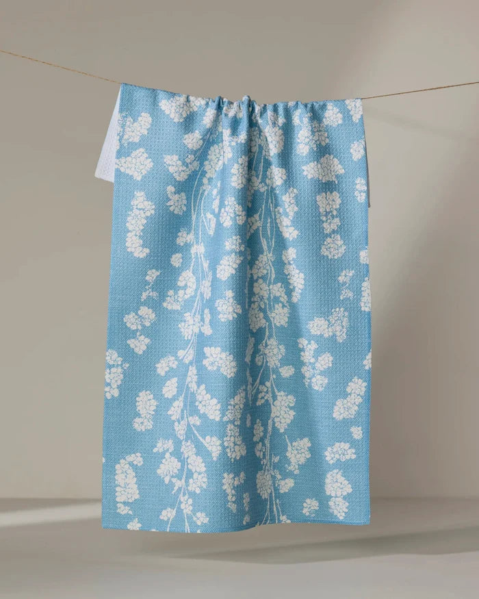 The Geometry Lilou Light Blue tea towel, featuring a white floral pattern, hangs neatly on a clothesline against a neutral background.