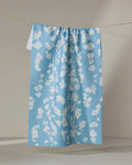 The Geometry Lilou Light Blue tea towel, featuring a white floral pattern, hangs neatly on a clothesline against a neutral background.