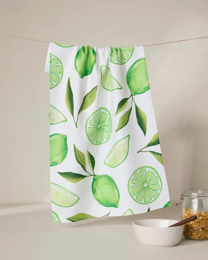 The GEOMETRY Lima kitchen towel, made from recycled materials and featuring green lime and leaf designs, hangs above a glass jar of brown sugar and a white bowl with a wooden spoon.
