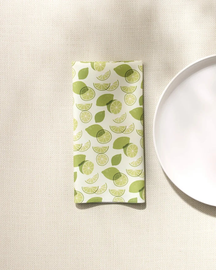 A folded GEOMETRY "Limes" eco-friendly napkin with a green lime fruit and slice design rests on a textured beige surface beside a white plate.