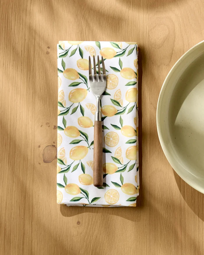 A fork rests on Geometry's Limoncello absorbent cloth napkin, featuring a yellow lemon pattern, atop a light wood table near a cream ceramic bowl. Sunlight creates soft shadows across the scene.