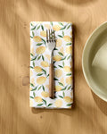 A fork rests on Geometry's Limoncello absorbent cloth napkin, featuring a yellow lemon pattern, atop a light wood table near a cream ceramic bowl. Sunlight creates soft shadows across the scene.