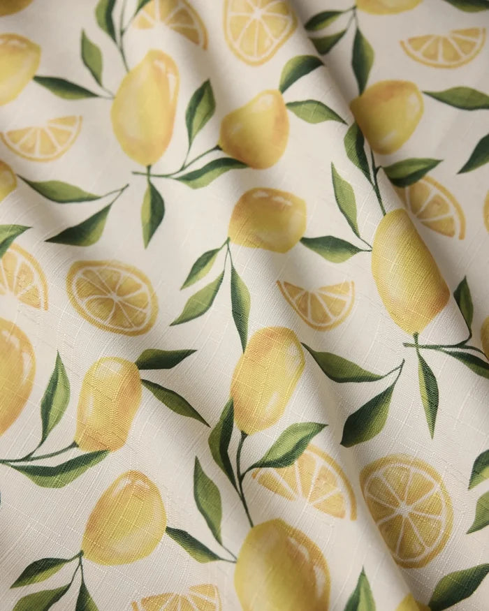 A close-up of Geometry’s Limoncello tablecloth shows a stain-resistant fabric with whole lemons, slices, and green leaves on a white textured background. Some wrinkles are visible.