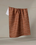 The Lined Mountains tea towel by Coit Creative features brown fabric with white wavy horizontal lines and hangs against a beige background, partly covering a white towel. This stylish kitchen accessory adds flair to any space.