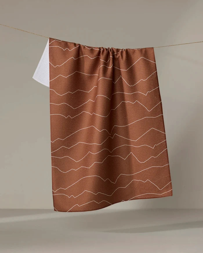 The Lined Mountains tea towel by Coit Creative features brown fabric with white wavy horizontal lines and hangs against a beige background, partly covering a white towel. This stylish kitchen accessory adds flair to any space.