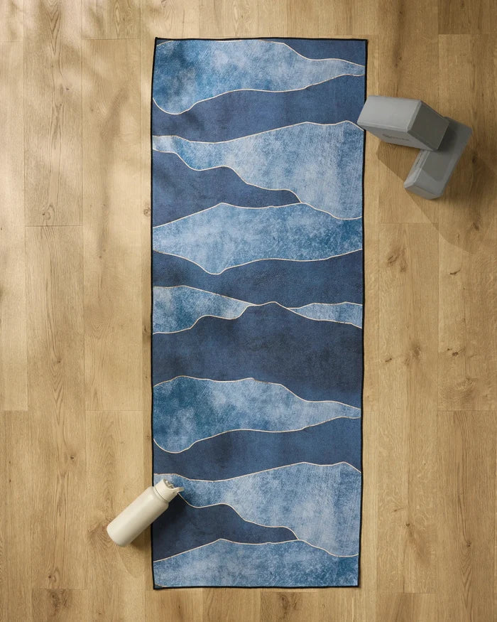 The GEOMETRY "Lined With Gold" yoga mat featuring blue with wavy white lines is spread on a wooden floor, topped with a slip-resistant towel. Two gray blocks are in the top right, and a white water bottle sits at the bottom left.