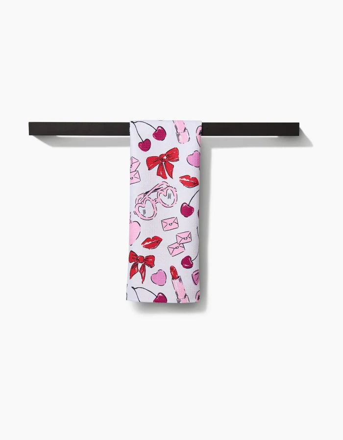 The Lip Smacking Good tea towel by Hufton Studio features pink and red hearts, bows, lips, envelopes, and perfume bottles on white fabric, hanging over a black bar against a white background.