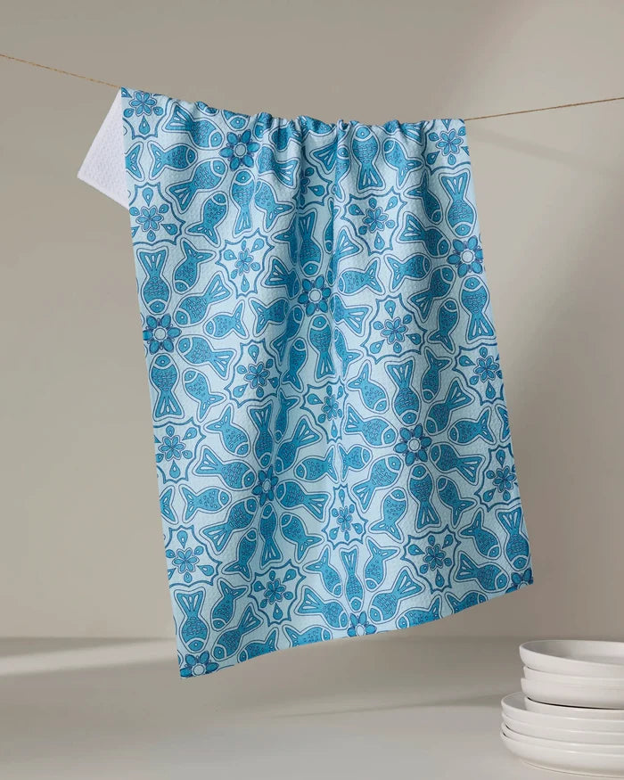 The Little Fish Mandala kitchen tea towel by Marta Harvey, made from recycled materials, features a blue and white fish and floral pattern. Shown on a clothesline beside white plates against a neutral background.