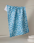 The Little Fish Mandala kitchen tea towel by Marta Harvey, made from recycled materials, features a blue and white fish and floral pattern. Shown on a clothesline beside white plates against a neutral background.