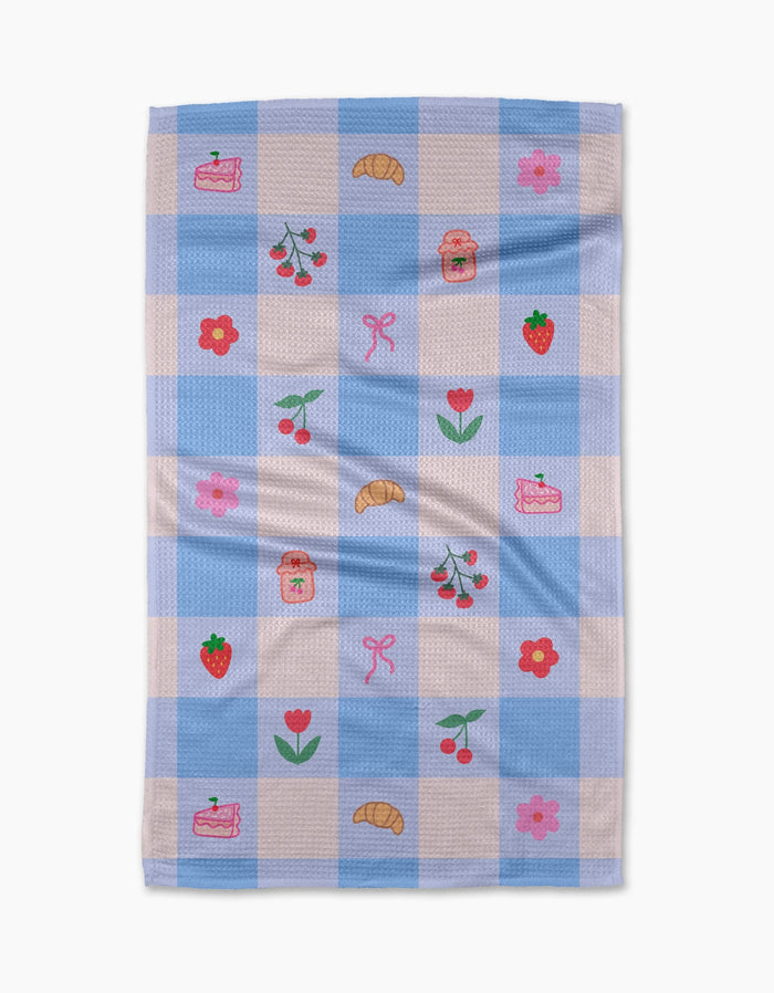 The Little Picnic Things tea towel by Letters By Shells is made from recycled materials and features blue and white checks with embroidered croissants, cherries, strawberries, jam jars, cake slices, pink bows, red tulips, and pink flowers.