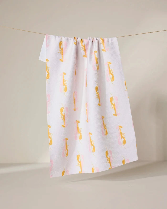 The GEOMETRY Lobster Love towel, made from recycled materials, features a white background with a yellow and pink abstract bird pattern and hangs on a clothesline against a neutral backdrop.