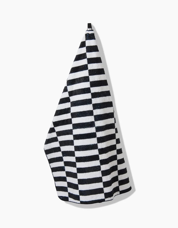 A Long Check towel by Hufton Studio, featuring black and white stripes and made from recycled materials, hangs from a hook against a plain white background, casting a shadow to the right.
