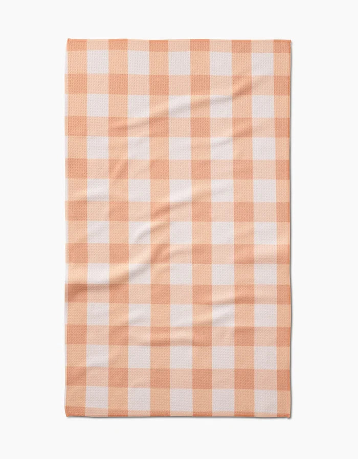 The Lorraine tea towel by Kelsi Fullmer is a rectangular Geoweave fabric towel made from recycled materials, shown flat with slight wrinkles and featuring a peach and white gingham checkered pattern.