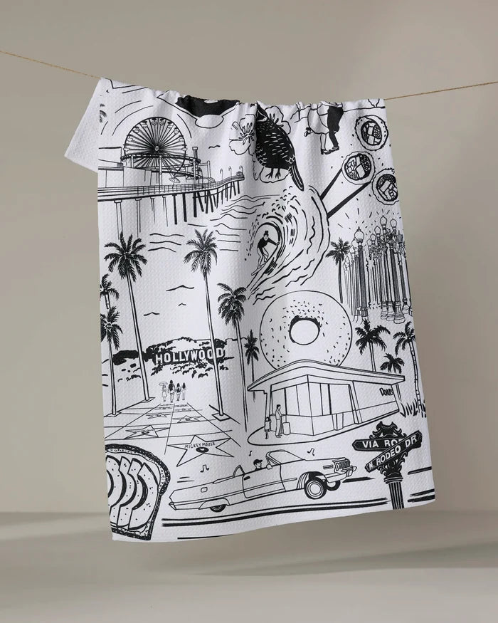 The Los Angeles kitchen towel by Elisabeth Wing is a white recycled towel with black line art, depicting iconic LA landmarks like the Hollywood sign, palm trees, a vintage car, donut shop, Ferris wheel, and street signs.