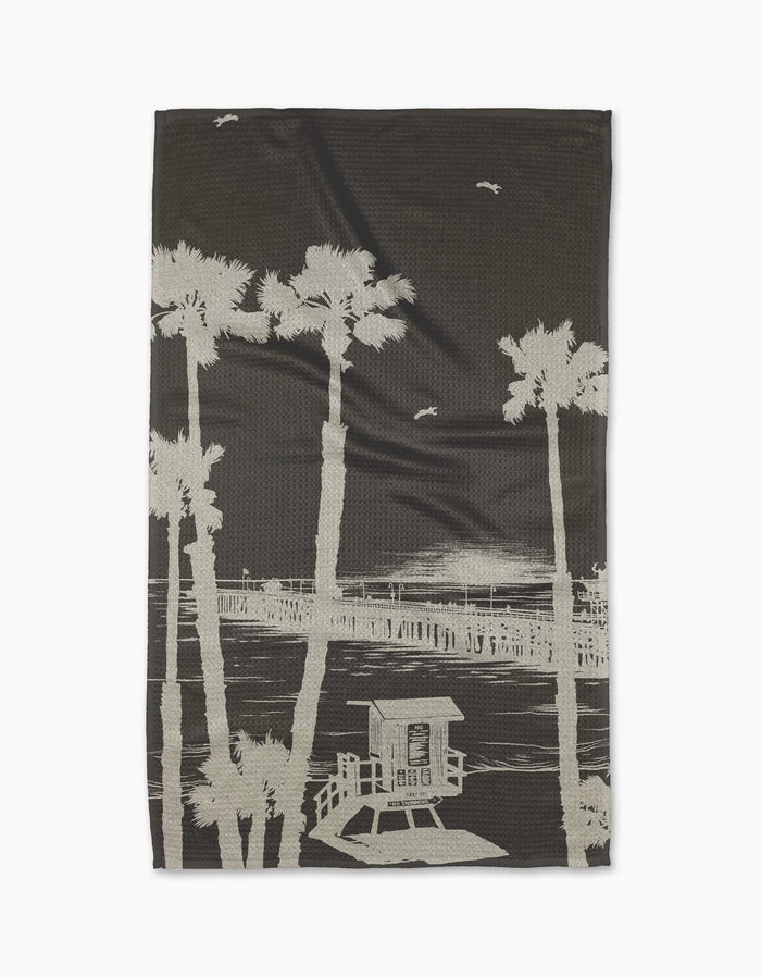 The Geometry Lost In Clemente blanket showcases a beige sketch of palm trees, a pier, and birds on a dark background, crafted from ultra-absorbent Geoweave fabric for comfort and style.
