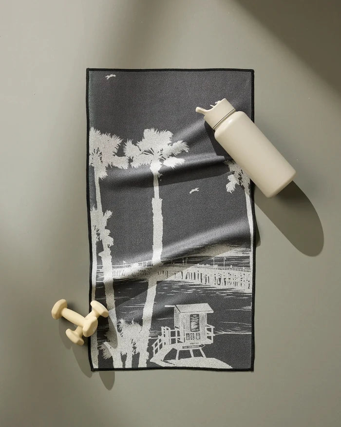 The Lost In Clemente fitness towel by Geometry, featuring a white beach and palm tree design on black, is ultra-absorbent and fast drying—perfect for your workout routine. Shown alongside an off-white water bottle and small dumbbells.